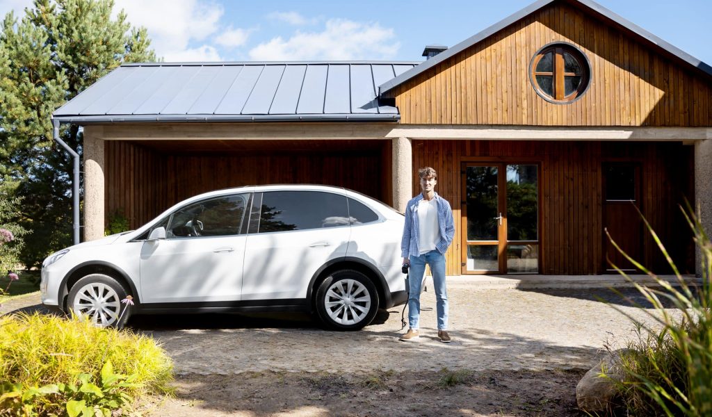 Homme en chemise ouverte devant son véhicule électrique blanc garé devant une maison à la façade en bois