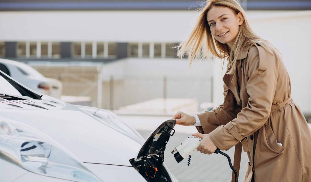 Femme en caban rechargeant son véhicule électrique blanc sur un parking d'entreprise